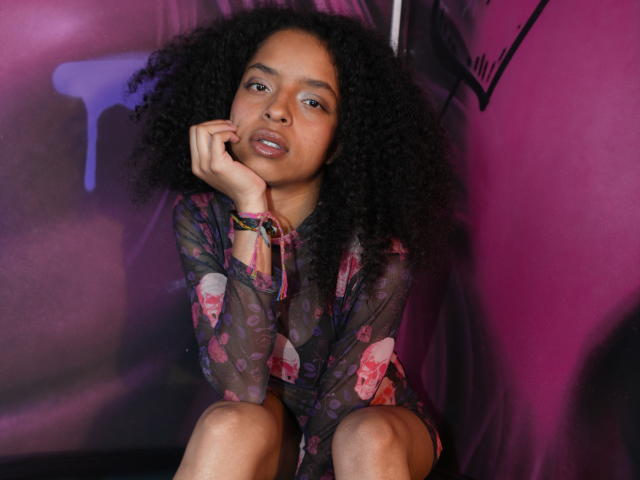 RihaLion, avec ses cheveux noirs et bouclés, se penche sur sa main, portant un haut transparent à motifs floraux, et est assise devant un fond rose-violet. Elle regarde séductrice l'appareil photo, avec la bouche légèrement entrouverte..
