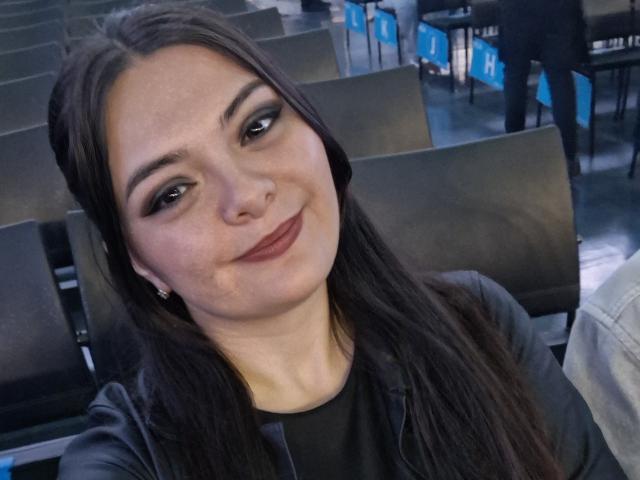 Selfie de perto de katherinLatin, sorrindo, com olhos castanho-escuros, delineador preto e batom. Ela tem cabelos longos e pretos, pele clara e veste uma blusa preta. Fundo iluminado em azul com cadeiras..