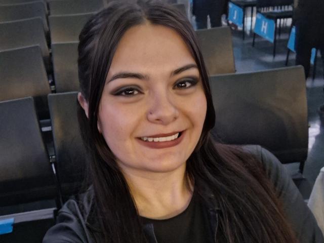 Gros plan de katherinLatin, une jeune femme aux longs cheveux noirs, souriant à l'appareil photo. Elle est assise dans un théâtre faiblement éclairé avec des sièges noirs. Ses yeux sombres et son maquillage discret attirent l'attention..