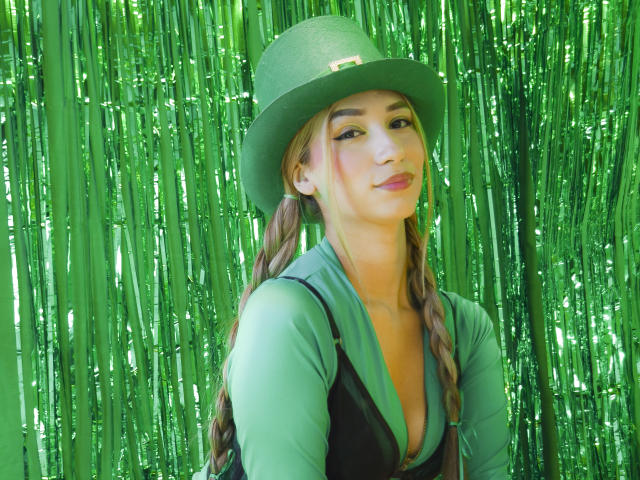 TanniaMoon, portant un chapeau vert et un haut noir, se tient devant des rideaux verts brillants. Elle a de longs cheveux tressés et une expression confiante, légèrement sensuelle. La lumière verte illumine son visage..