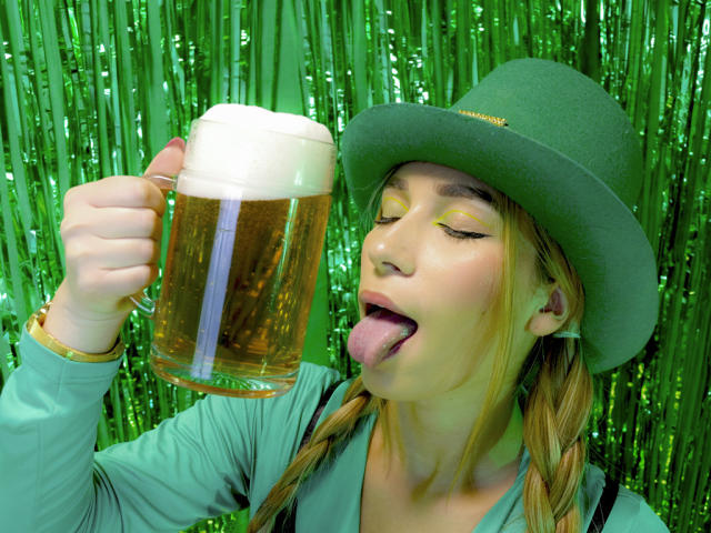 TanniaMoon con gli occhi chiusi e la lingua fuori, che tiene una pinta di birra schiumosa nella mano destra. Indossa un cappello verde e una camicia verde, con uno sfondo di paillettes verdi..