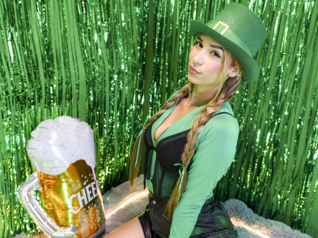 TanniaMoon met lang gevlochten haar, een groene hoed en een zwarte jurk, die een schuimend biertje vasthoudt met het label "CHEER" erop. glanzend groen gordijn.
sexy, biertje in de voorgrond, haar blik is verleidelijk..