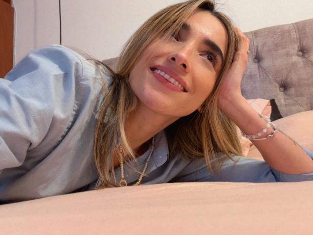 AdharaCooperr lying on a bed, smiling, head resting on hand, wearing a light blue shirt, brown hair, casual pose, soft lighting, tufted headboard, white pillow, relaxed expression.