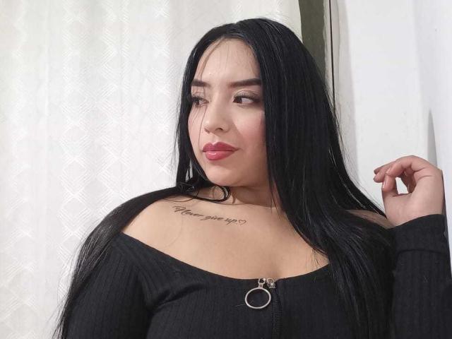 Photograph of AdaraCute69 with long black hair, wearing a black off-shoulder top, red lipstick, and a tattoo on her chest. She's gazing sideways with a finger in her hair. Fucking sexy.