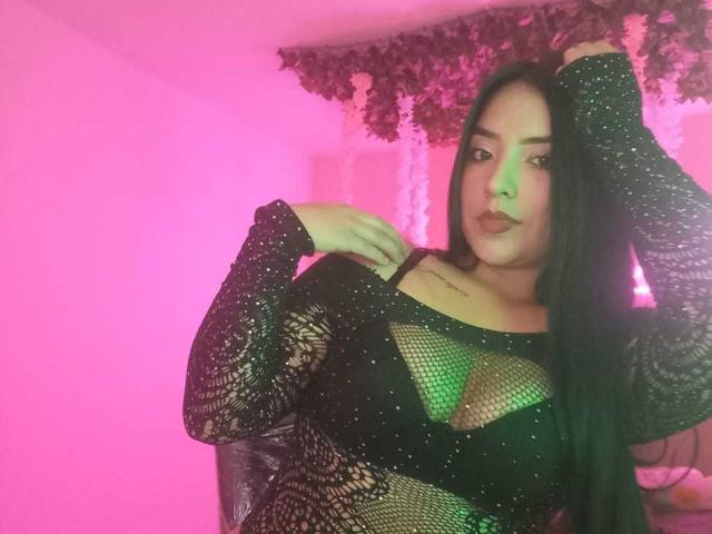 AdaraCute69, une femme séduisante avec de longs cheveux noirs, porte un haut en maille noire scintillant qui révèle un soutien-gorge vert, et pose de manière suggestive. Avec une main sur sa tête. Fond rose vif.