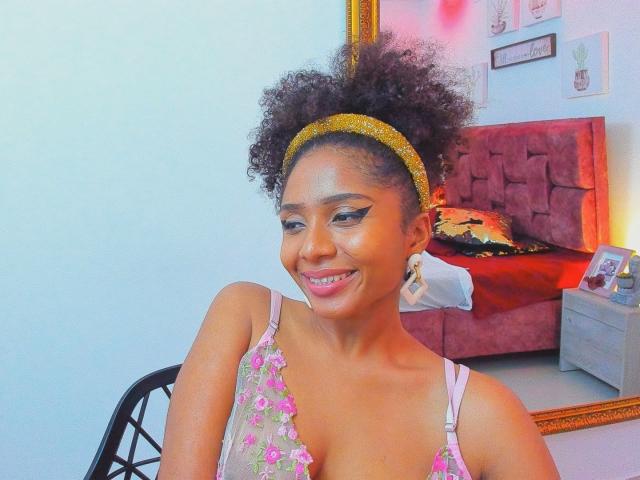 Megan Williams, met donkere huid en krullend haar, draagt een gele haarband en een topje met bloemenprint, en glimlacht naar de camera. De achtergrond toont een rood bed met een gloeiende lamp. Er is duidelijke seksuele spanning aanwezig..