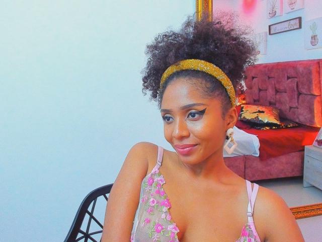 Fotografia di Megan Williams, una donna afroamericana attraente con i capelli ricci, che indossa una fascia dorata e una camicetta rosa con fiori. Seduto in una stanza con un divano rosso e pareti bianche. Molto attraente..