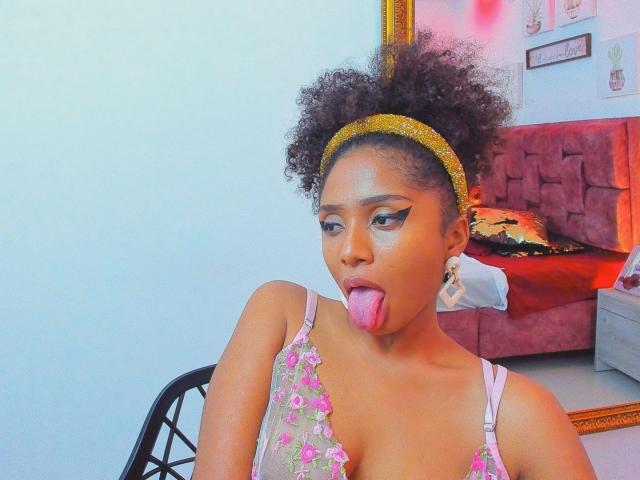 Photograph of MeganWilliams with dark skin, curly hair, yellow headband, tongue out, floral tank top, earrings, seated in room with red couch, white wall, and framed picture. Fucking sexy look.