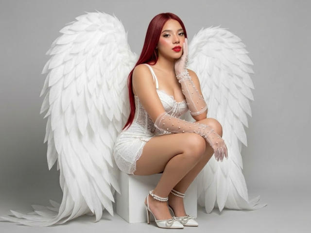 MiaLeroy está sentada en un pedestal blanco, vestida con un vestido blanco de encaje y tacones altos blancos, y lleva grandes alas blancas de ángel. Ella tiene el pelo largo y rojo, y lápiz labial rojo, luciendo seductora..