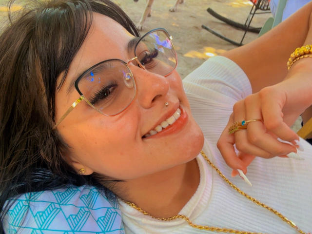 Close-up photo of GraceClarke with light brown skin, black hair, and round glasses, smiling while lying down. She wears a white top, gold necklace, and rings, with a relaxed, sexy vibe.
