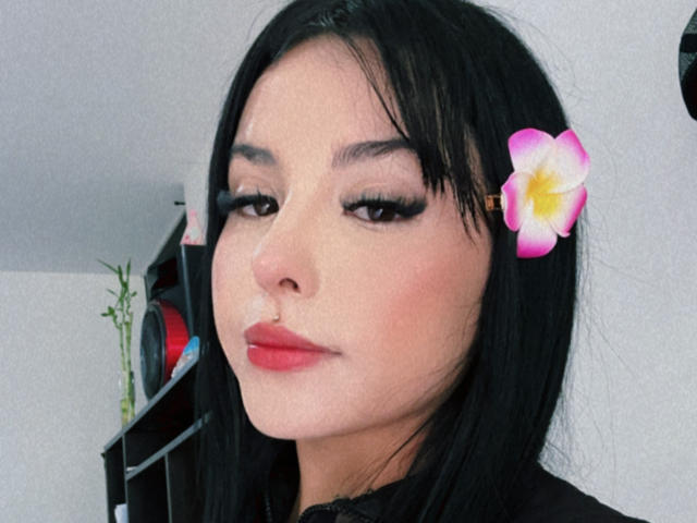 Close-up of GraceClarke, a sexy Asian woman with black hair, red lipstick, and a pink flower in her hair. Her eyelids are heavy with black mascara, creating a seductive look.