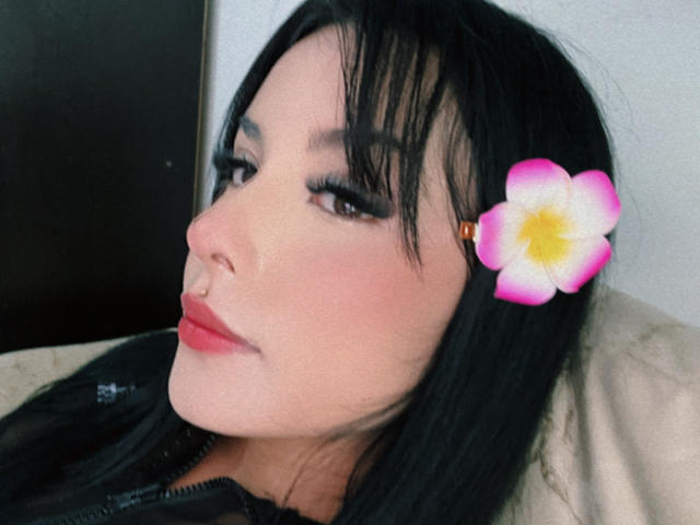 Close-up of GraceClarke, an Asian woman with black hair, red lipstick, and a pink flower clip, looking slightly to the side. Her long eyelashes are prominent, and her expression is sultry.
