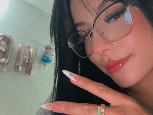 Close-up of GraceClarke, wearing clear glasses, red lipstick, and long, pointed nails with a gold ring, touching her face with one hand, showing off her cock ring on her finger.