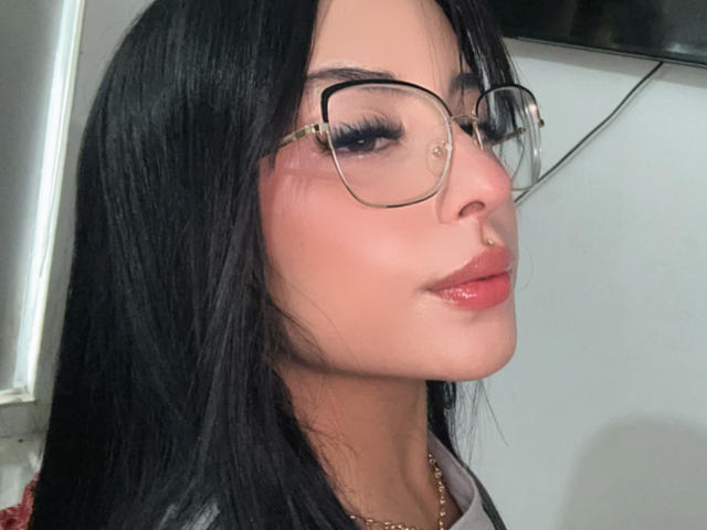 Primer plano de GraceClarke, una mujer con cabello largo y negro, que lleva gafas redondas y lápiz labial rojo. Tiene una ligera sonrisa, labios carnosos y está mirando hacia un lado. El fondo está desenfocado..
