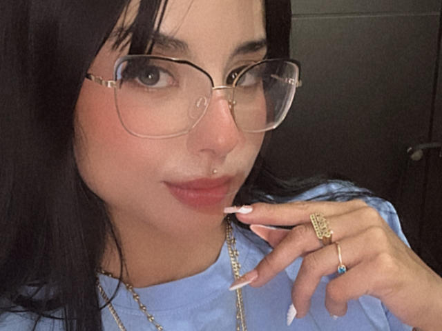 Close-up selfie of GraceClarke, with large glasses, black hair, and light blue shirt. She's biting her manicured finger, wearing a gold ring and necklace. Her expression is sultry.