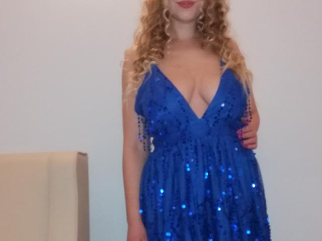 ChgarminitaHot in a low-cut, blue sequined dress, showing ample cleavage. She has long, curly blonde hair, fair skin, and her right hand resting on her hip. Seductive, sexy pose.