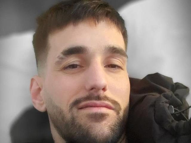 Foto em close-up de Moielle69, um homem de barba com cabelo castanho curto, olhos ligeiramente semicerrados e um sorriso discreto. Sombras suaves em seu rosto, vestindo uma jaqueta preta. Tensão sexual..