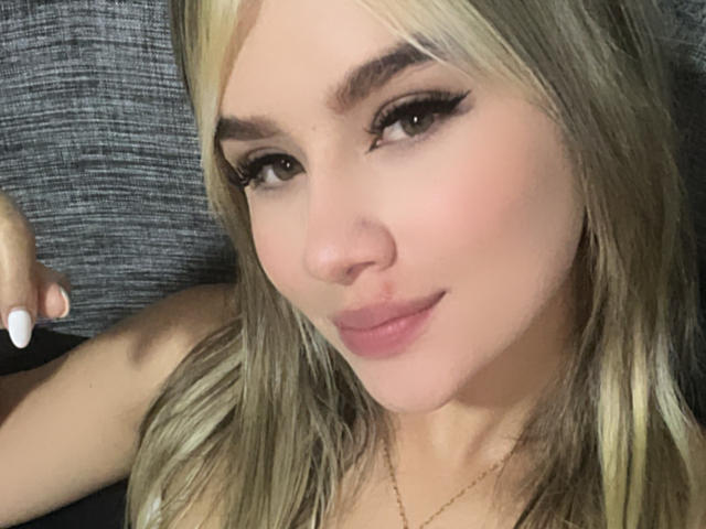 Close-up selfie of SashaPolet, blonde hair, heavy black eyeliner, full lips, fair skin, gold necklace, right arm raised, gray textured background. Slight smirk, soft lighting, slight shadow on face.