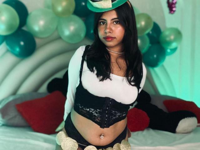 Fotografía de Celeste Morel, de pie frente a una cama con globos verdes y turquesa. Ella lleva una camiseta blanca corta, un corpiño de encaje negro, pantalones cortos negros y un sombrero verde. Pelo oscuro, muy atractivo, con piercing en el ombligo..