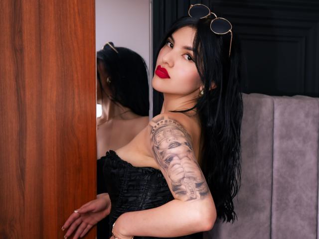 Photograph of MonseBelucci, with long black hair, red lipstick, and a tattooed arm, leaning against a wooden door, wearing a black strapless dress, eyeglasses on head, sultry expression.