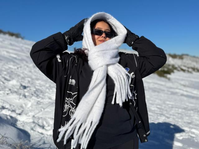 Sophia Browm está em uma paisagem coberta de neve, vestindo uma jaqueta preta com padrões brancos, um cachecol branco e luvas pretas. Ajustando o capuz com as mãos. Ela está usando óculos de sol escuros, com um céu azul claro ao fundo. Que frio!.