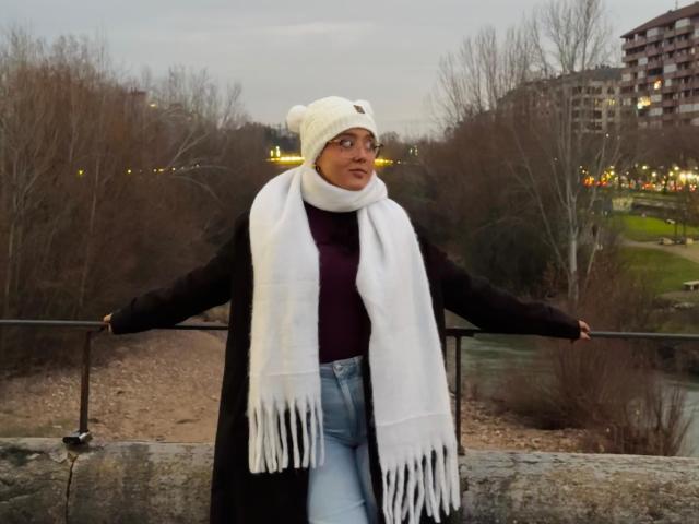 SophiaBrowm stands leaning against a railing, wearing a white beanie, white scarf, black coat, and blue jeans. She's gazing to the right, with urban buildings and trees in the background. Fucking cold look.