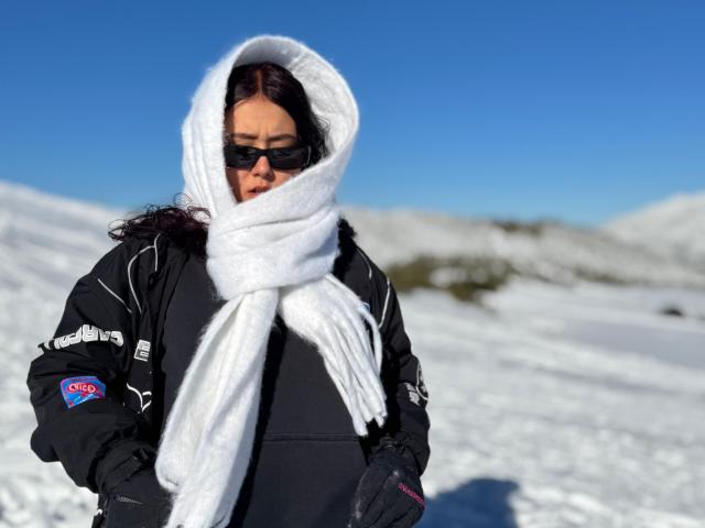 Foto van Sophia Browm met donker haar, die een witte bontvoering met capuchon, een zwarte jas en zwarte handschoenen draagt. Ze draagt een zonnebril en staat in een besneeuwd landschap met een blauwe lucht. Vreselijk koud..