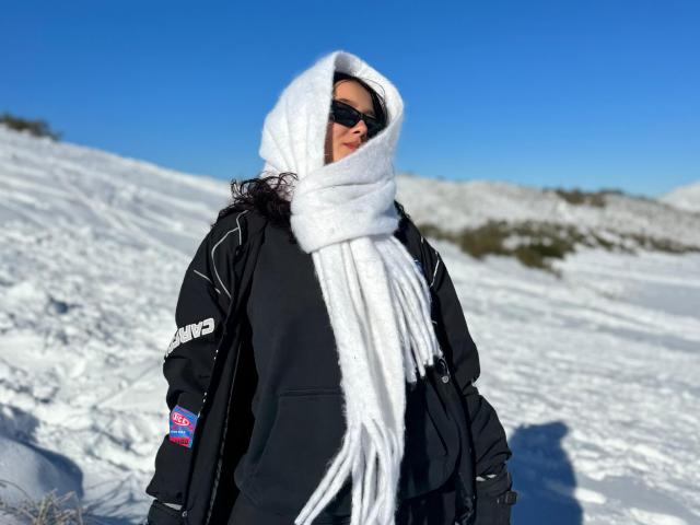 Sophia Browm está de pie en un paisaje nevado, vistiendo una chaqueta negra con detalles blancos, una bufanda blanca gruesa y oscura. gafas de sol. Cielo azul brillante y colinas cubiertas de nieve al fondo. Hace un frío que da asco..
