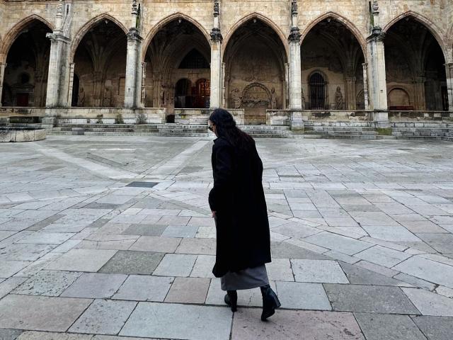 Una figura solitaria, SophiaBrowm, camina a través de un amplio y vacío patio de piedra con arcos en el fondo. Sophia Browm lleva un abrigo largo negro y botas, proyectando una sombra en el suelo..