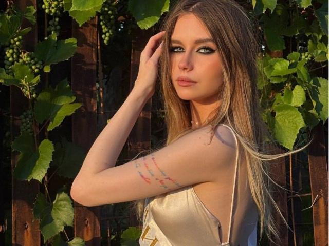 Fotografie von Librada Pylvainen, die mit leicht gewandtem Rücken steht, eine Hand in ihrem braunen Haar, und ein ärmelloses Kleid trägt. Goldenes Kleid mit Tattoos auf ihrem rechten Arm. Von der Sonne durchflutet, umgeben von grünen Ranken. Verdammt sexy..