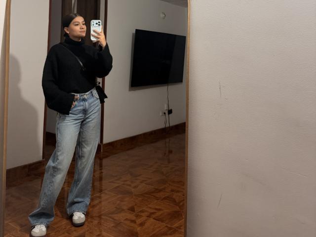 ThaliaAres steht vor einem Spiegel und hält ein Handy mit einer schwarzen Hülle, um ein Selfie zu machen. Sie trägt eine schwarze Kapuzenjacke, blaue Jeans und weiße Turnschuhe. Verdammt heiß. Die Spiegelung zeigt einen Flachbildfernseher..