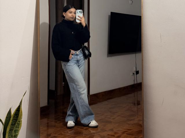 ThaliaAres, een vrouw met kort haar, staat in een kamer en maakt een spiegelzelfportret. Ze draagt een zwarte top, lichtblauwe jeans en witte sneakers, en ze houdt een telefoon met een zwarte hoes vast..