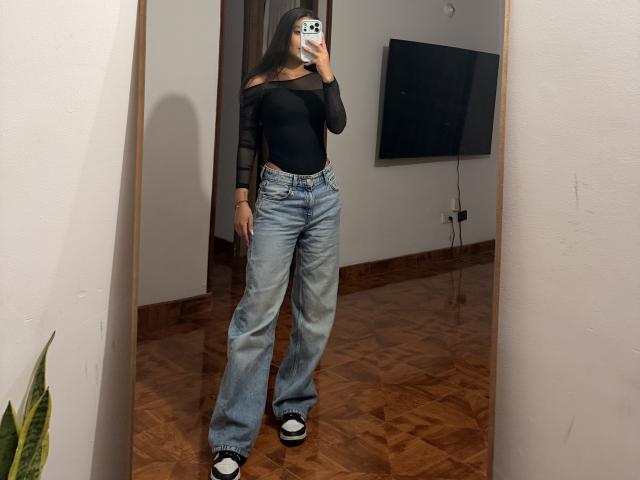 ThaliaAres se tient devant un miroir, vêtue d'un haut noir à une épaule et d'un jean bleu ample, tenant un téléphone devant elle. de son visage. Ceinture en flanelle et chaussettes blanches. Pièce avec parquet..