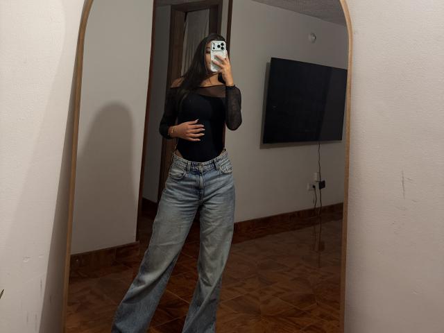 ThaliaAres si trova in una stanza poco illuminata e sta scattando un selfie allo specchio. Indossa una maglia nera trasparente, jeans blu a vita alta e tiene il telefono vicino al viso. Ombre proiettate sulla parete.