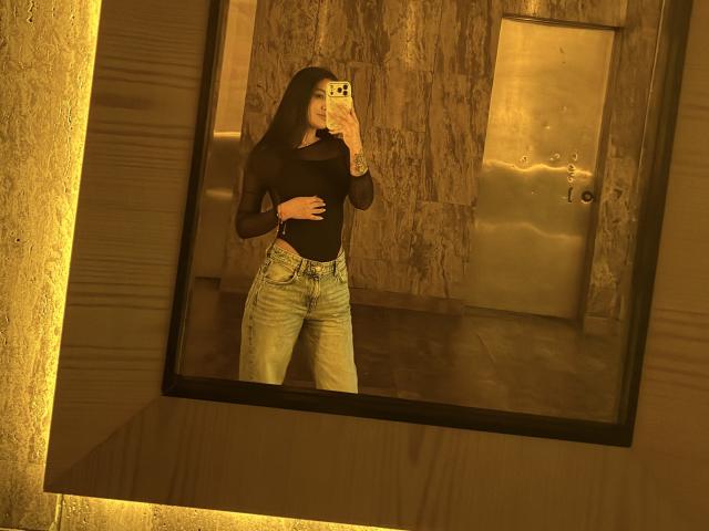 ThaliaAres, um reflexo em um espelho, vestindo uma blusa de manga comprida preta e jeans azul claro, segurando um telefone para tirar uma foto. selfie, sexy, iluminação em tons de amarelo dourado, parede de mármore ao fundo.