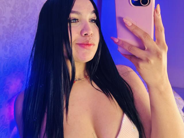 MiaDenver, een vrouw met bleke huid en lang zwart haar, maakt een selfie met een roze telefoon. Ze draagt een witte top met een lage halslijn, waardoor veel decolleté zichtbaar is. Haar uitdrukking is subtiel verleidelijk. Blauw licht schijnt achter haar..