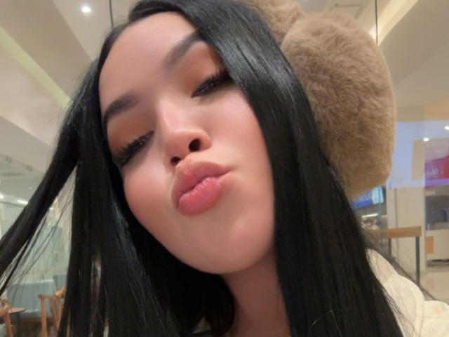 Close-up photo of MiaDenver, a hot Asian woman with long black hair, puffy lips, and a furry hat. Her eyes are half-closed, giving a sultry, sexy look. Her cock is not visible.