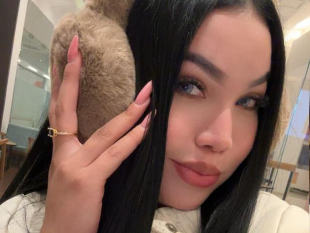 Close-up of MiaDenver with long black hair, blue eyes, and red lipstick, holding a brown fur collar against her face. She wears a gold ring on her left hand, with red nails. Fucking sexy.
