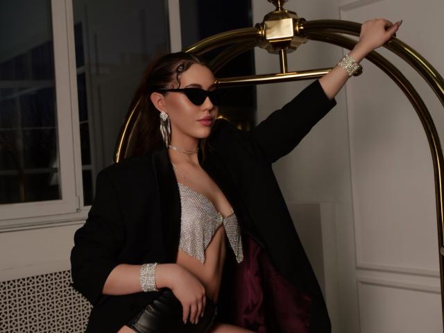 BloomAndFun se apoya en una lámpara de pie de latón, vistiendo un abrigo negro sobre una blusa brillante y escotada, y gafas de sol negras. Joyería de diseño llamativo y con formas grandes. Los dedos tocan la lámpara, irradiando un atractivo seguro..