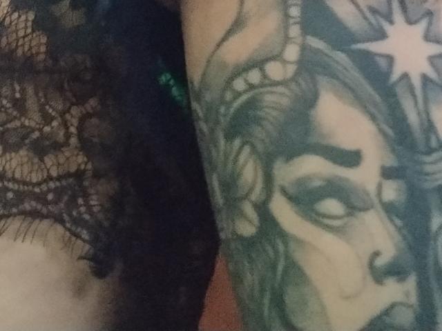Gros plan du bras de GrethelMulher, présentant un tatouage noir et vert détaillé représentant un visage féminin serein avec une étoile. Fond noir.
sa lingerie noire ornée de motifs en dentelle est visible sur la gauche..