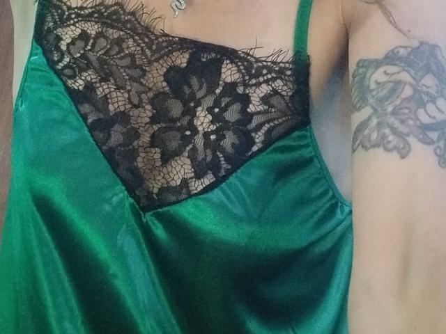 Nahaufnahme des Oberkörpers von GrethelMulher, die ein grünes Seidenkleid mit schwarzer Spitzenüberlagerung trägt. Ihr Arm, verziert mit einem detaillierten floralen Tattoo, ruht an ihrer Seite und gibt einen Hauch von Dekolleté preis..