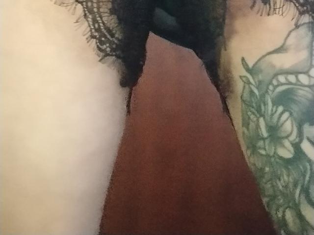 Close-up of GrethelMulher's hip and upper thigh, showing a black lace garter on the left and a detailed green and black tattoo on the right, with a reddish-brown background.