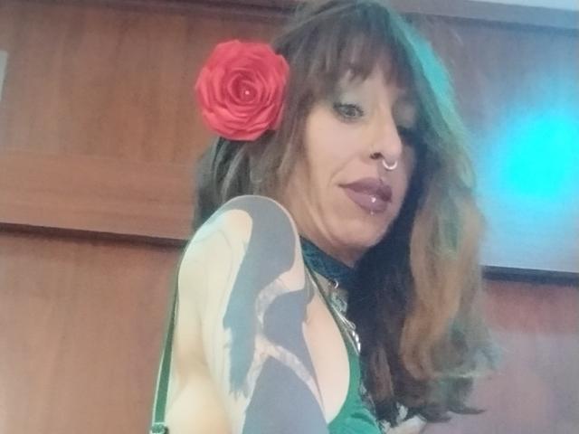 Fotografía de GrethelMulher, una mujer de piel marrón media, con cabello largo y ondulado, una rosa roja en el cabello, lápiz labial morado, nariz. anillo, vistiendo una blusa negra, luz azul a la derecha, fondo de madera, tensión sexual.