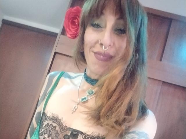 Close-up of GrethelMulher, brown-haired woman with a red rose in her hair, nose ring, and dark lipstick, wearing a lace-patterned top, teal choker, and blue-green fabric. Fucking sexy smile.