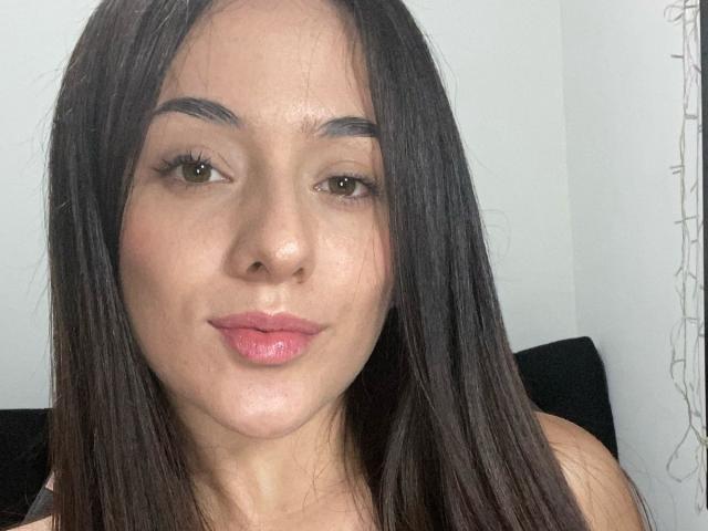 Close-up of NoahNaughty with straight, dark brown hair, light skin, and pink lips. She has arched eyebrows and is looking directly at the camera with a subtle, confident smile.