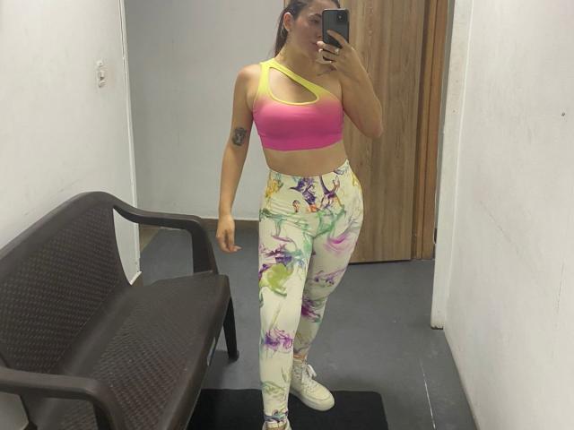 NoahNaughty staat in een helder verlichte kamer en maakt een spiegelzelfportret. Een roze sportbeha, kleurrijke leggings met bloemenprint en witte sneakers. Zichtbare tatoeage op de rechterarm, zwarte stoel in de buurt..