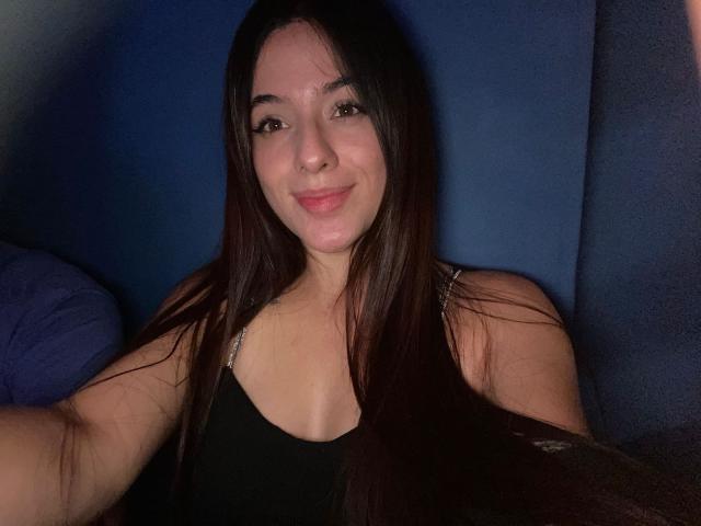 Fotografía de NoahNaughty, una mujer sonriente con cabello largo y oscuro, vestida con una camiseta negra sin mangas, sentada contra un fondo azul. Antecedentes. Su piel está radiante, los ojos ligeramente entrecerrados, lo que crea un ambiente sensual..