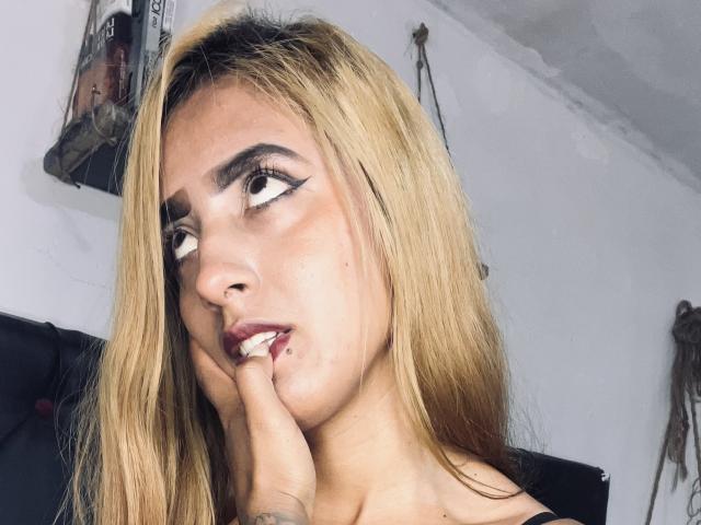 Close-up of NicolForest, a blonde woman with heavy black eyeliner, biting her finger with a sultry expression. Bright light highlights her face, creating shadows on her cheek and neck.