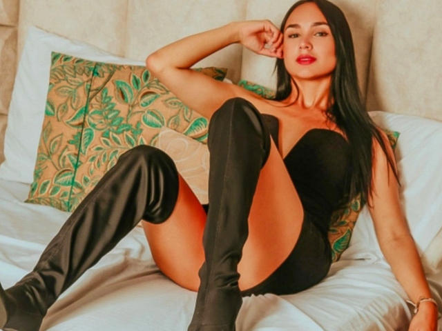 LunaMaxwell ligt op een bed, met gespreide benen, en draagt een zwart, mouwloos body en kniehoge laarzen. Haar lange zwarte haar, rode lippen en zelfverzekerde houding stralen een sensuele spanning uit. Bloemenkussen op de achtergrond.