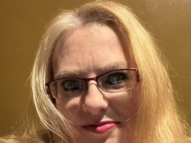 Foto ravvicinata di Cherish Williams, una donna bionda con la pelle chiara, che indossa occhiali con montatura rossa e rossetto rosa, con un'espressione intensa. sguardo fisso, occhi leggermente socchiusi, sfondo marrone dorato, la luce solare intensa illumina i capelli e il viso..